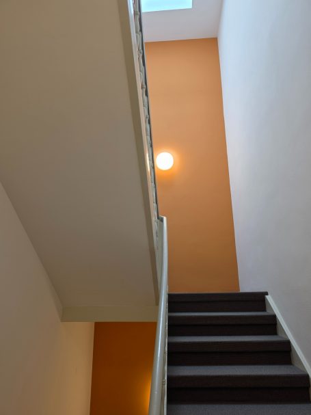 Treppenaufgang mit oranger Wand und einer Deckenlampe im oberen Bereich.